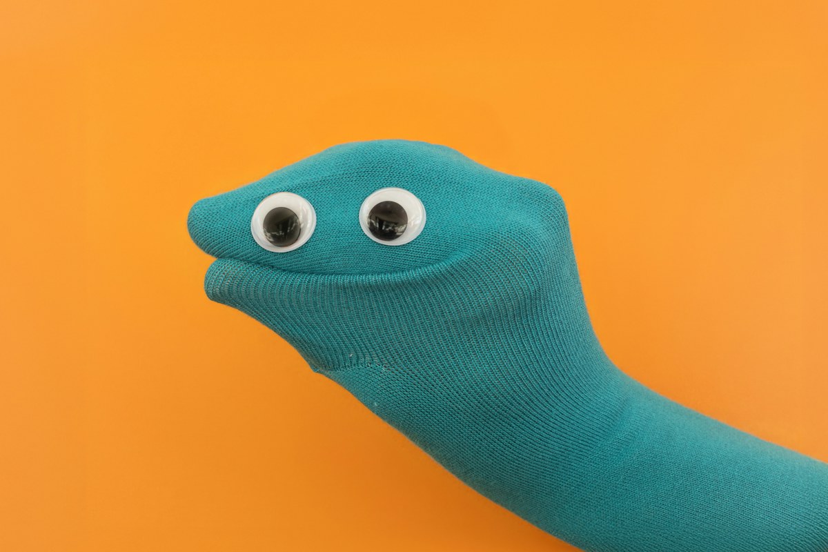 A turquoise sock puppet with large plastic eyes appears against an orange background. Illustrative photo. This activity uses drawn-on features rather than small loose decorations.