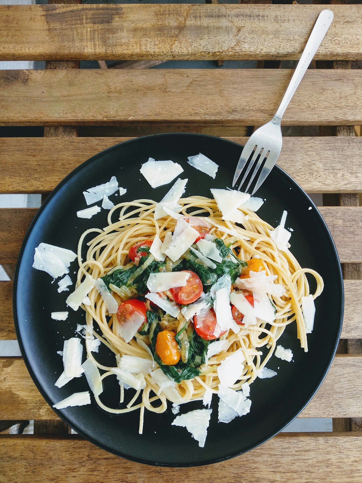 Spaghetti with tomatoes, spinach, and shaved cheese on a dark plate. Illustrative food photograph; the finished recipe may look different.