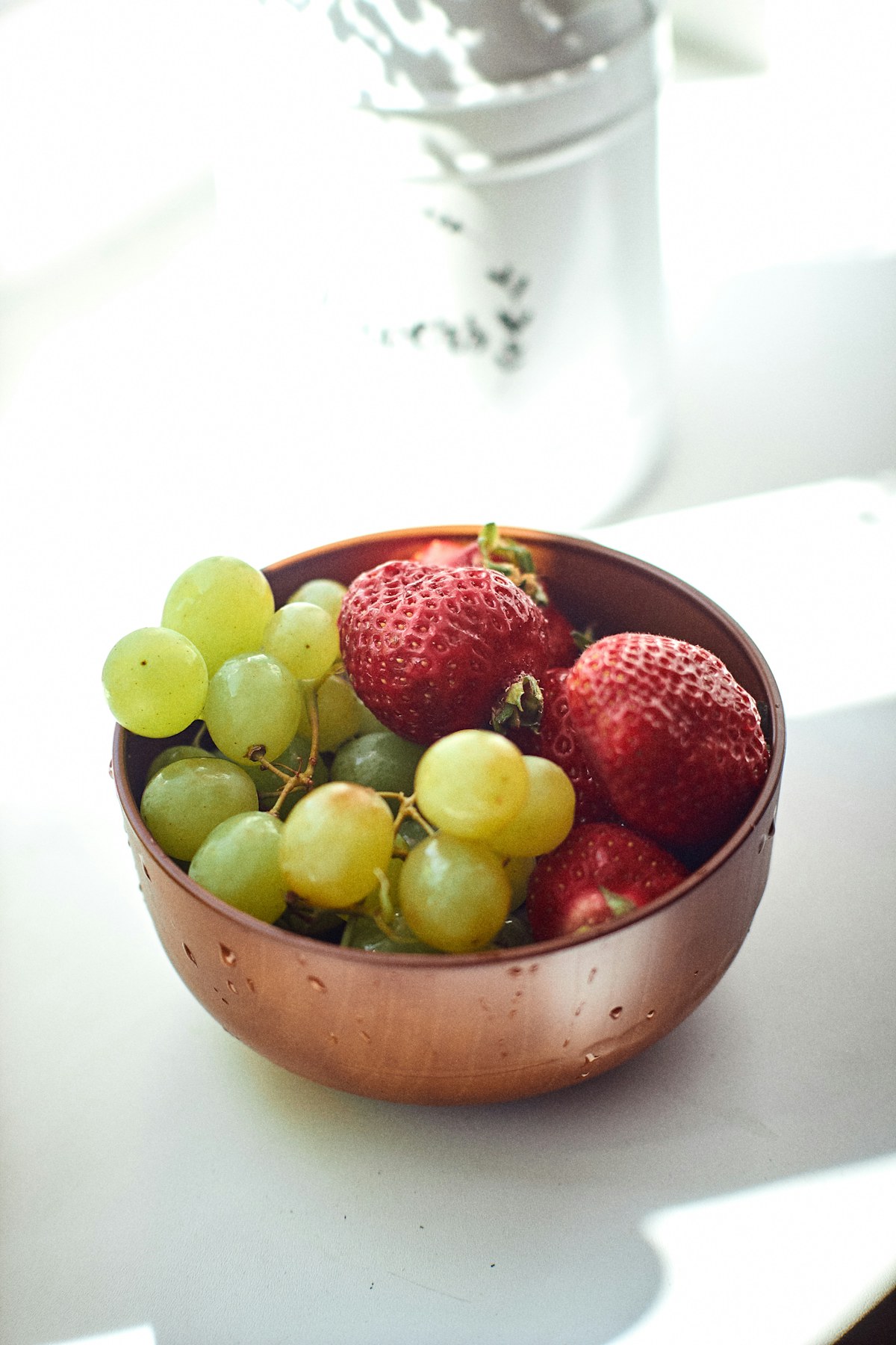 Fresh strawberries and green grapes in a copper bowl on a white counter. Illustrative photograph for an original fictional story.