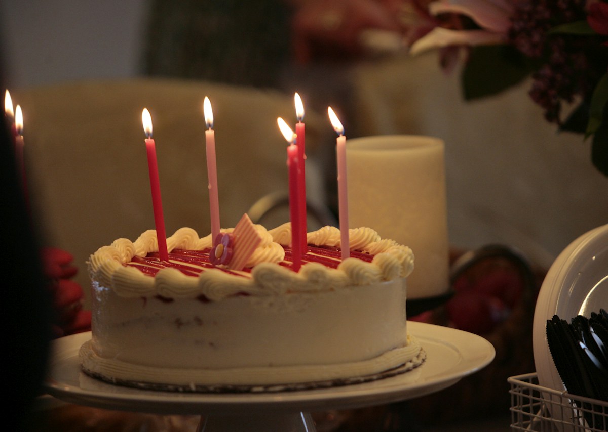 A white birthday cake with red decorations and lit candles on a wooden table. Illustrative photograph for an original fictional story.